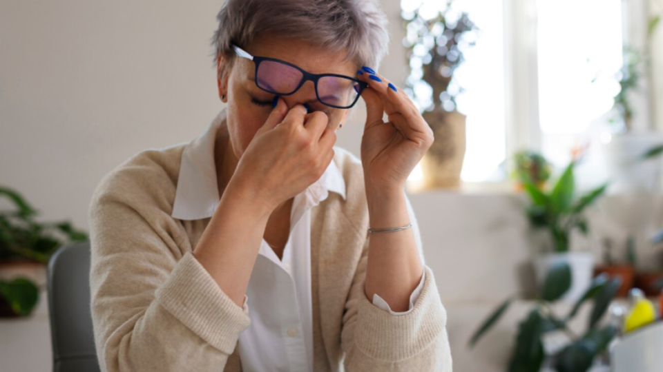 Retrata uma mulher de cabelos curtos grisalhos sentada em uma mesa de escritório ou home office, rodeada por plantas ao fundo. Ela levanta levemente seus óculos de armação escura com uma das mãos enquanto pressiona os olhos fechados com a outra, simbolizando cansaço visual após longas horas de leitura ou trabalho no computador.