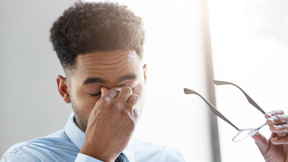 Apresenta o plano aproximado de um homem de camisa social azul e gravata que removeu seus óculos de grau (segurados com a mão esquerda) para massagear a ponte do nariz com os olhos fechados. A imagem transmite de forma nítida os conceitos de fadiga ocular, estresse profissional ou forte dor de cabeça.