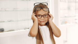 Uma menina jovem com longos cabelos castanho-claros está em uma ótica, sorrindo timidamente enquanto experimenta e empilha vários pares de óculos na cabeça.