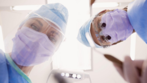 A imagem mostra dois profissionais de saúde, provavelmente dentistas ou cirurgiões, fotografados de um ângulo inferior (perspectiva do paciente deitado).